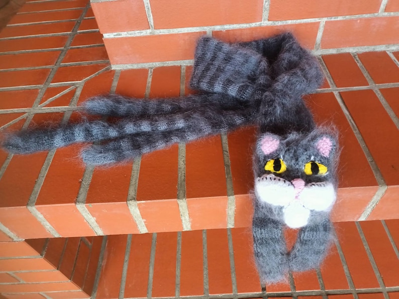 May include: A gray and black striped knitted scarf with a cat face at the end. The cat face has pink ears, yellow eyes, and a white nose and mouth.