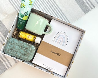 May include: A white gift box filled with a variety of items, including a green mug, a bar of soap, a bottle of oil, a tube of hand cream, and a card with a rainbow design and the text "Zaubervilla" and "DATUM".