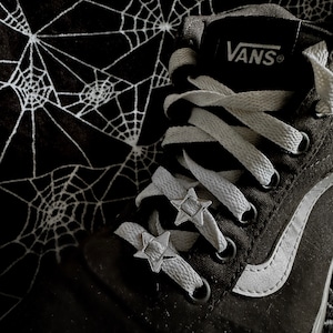 Puede incluir: Zapatillas Vans negras con cordones blancos y una franja blanca en el lateral. Los cordones están decorados con abalorios en forma de estrella plateados. Las zapatillas están sobre un fondo con estampado de telaraña negra y blanca.