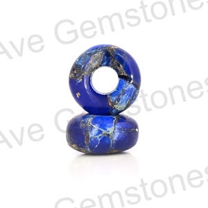 Puede incluir: Dos cuentas de donut de piedra preciosa azul con apariencia veteada. Las cuentas tienen un agujero en el centro y están apiladas una encima de la otra. Las cuentas tienen un acabado pulido y son de un azul intenso con vetas doradas y blancas.