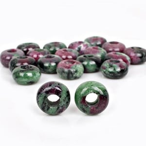 Puede incluir: Un grupo de cuentas de piedra preciosa verde y rosa con un agujero en el centro. Las cuentas están dispuestas en un montón suelto sobre una superficie blanca. Dos cuentas están separadas del montón y están en foco.