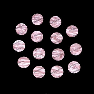 May include: A close-up of a collection of 12 round, pink, iridescent, decorative, gemstone-like objects. The objects are arranged in a circle on a black background. The text "Ave Gemstones" is printed on the objects.