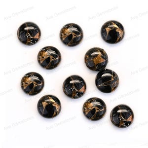 Könnte beinhalten: Zwölf runde schwarze Cabochons mit goldenen Flocken. Die Cabochons sind auf einer weißen Oberfläche verstreut.