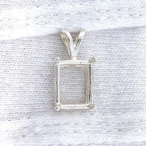 Puede incluir: Un engaste de colgante plateado con forma rectangular, diseñado para sostener una piedra preciosa. El engaste tiene un lazo en la parte superior para una cadena y pequeñas cuentas decorativas en cada esquina. El colgante se muestra sobre una superficie blanca texturizada.