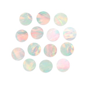 May include: A collection of 13 iridescent opal cabochons, each with a unique pattern of colors and a slightly different shape. The cabochons are arranged in a circle on a white background. Ave Gemstone is printed on some of the cabochons.
