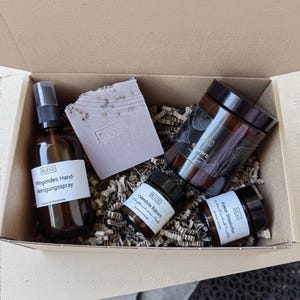 Op de afbeelding: Een geschenkdoos met een bruine glazen sprayfles met het label Pflegendes Hand-Reinigungsspray, een rechthoekig lichtpaars stuk zeep met een BLES2-stempel, een grote bruine glazen pot met het label flowers en twee kleine bruine glazen potjes met de labels Calendula Balsam en Rosen Shaebister.