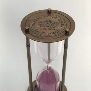 May include: Brass hourglass with a pink sand timer. The top of the hourglass has a circular brass plate with the inscription "Makers to the Queen London Sand Timer".