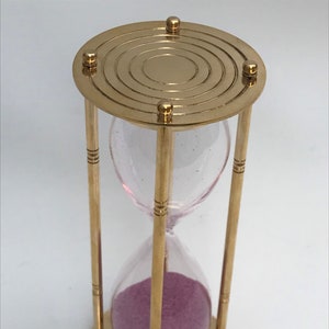 May include: A brass hourglass with a pink sand timer. The hourglass has a circular top with concentric circles and a base with a similar design.