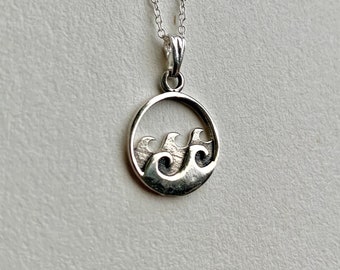 May include: A silver necklace with a circular pendant featuring a wave design. The pendant is detailed with three waves and a textured surface.
