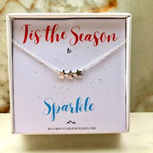 Könnte beinhalten: Eine silberne Halskette mit drei goldenen Sternanhängern. Die Halskette befindet sich in einer weißen Schachtel mit dem Text "Tis the Season to Sparkle" und der Website "bluemountainjewellery.com".