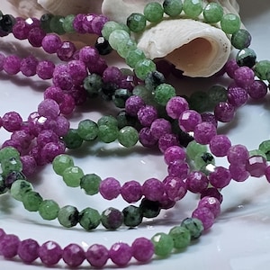 Può includere: Un filo di perline rotonde sfaccettate caratterizzato da un mix di pietre rosa magenta vibrante e verde chiaro con occasionali inclusioni nere. Le perline sono infilate in una linea continua, mostrando una consistenza lucida e un motivo cromatico variegato.