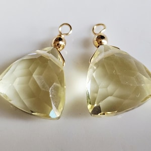 Op de afbeelding: Twee gefacetteerde, lichtgele edelsteenhangers met goudkleurige metalen beslagen. De edelstenen zijn driehoekig met meerdere facetten, die het licht vangen. De hangers worden op een witte ondergrond getoond.