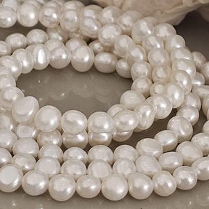 Op de afbeelding: Close-up van een streng glanzende, witte parels. De parels zijn onregelmatig van vorm, met een glad, reflecterend oppervlak. De parels zijn aan elkaar geregen en vormen een ketting of armband. Het thema is sieraden en mode.