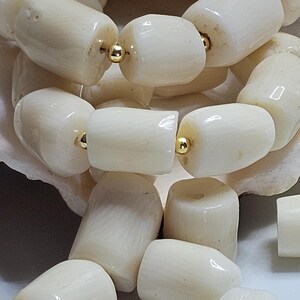May include: Close-up of a cream-colored beaded necklace. The beads are cylindrical and strung together with gold-colored metal. The necklace is resting on a white surface.