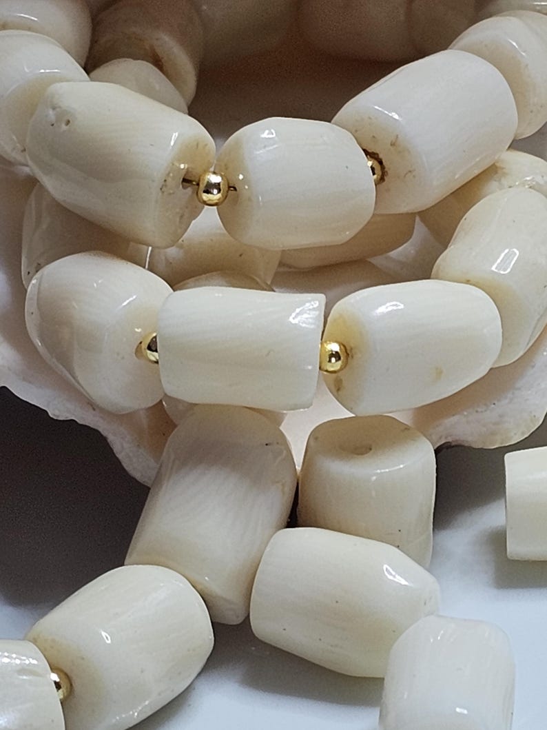 May include: Close-up of a cream-colored beaded necklace. The beads are cylindrical and strung together with gold-colored metal. The necklace is resting on a white surface.