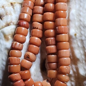 Op de afbeelding: Close-up van drie strengen koraalkralen. De kralen hebben een warme oranjerode kleur en zijn cilindrisch van vorm. De kralen zijn aan elkaar geregen, waardoor een ketting of armband ontstaat. De achtergrond is een licht, getextureerd oppervlak.