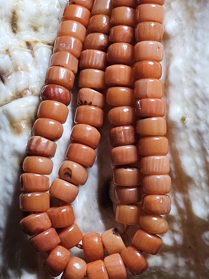 Op de afbeelding: Close-up van drie strengen koraalkralen. De kralen hebben een warme oranjerode kleur en zijn cilindrisch van vorm. De kralen zijn aan elkaar geregen, waardoor een ketting of armband ontstaat. De achtergrond is een licht, getextureerd oppervlak.