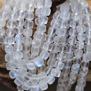 May include: A strand of white moonstone beads, each bead is faceted and has a square shape. The beads are strung on a clear thread and are arranged in a loose pile.