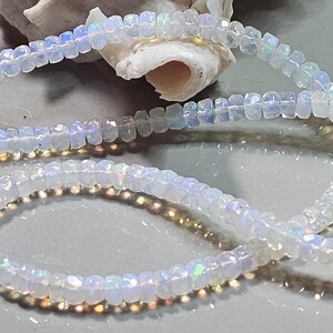 May include: A strand of small, faceted, translucent white opal beads with iridescent flashes of blue, green, and yellow light. The beads are circular and strung together in a continuous line, displaying a reflective, polished surface texture.