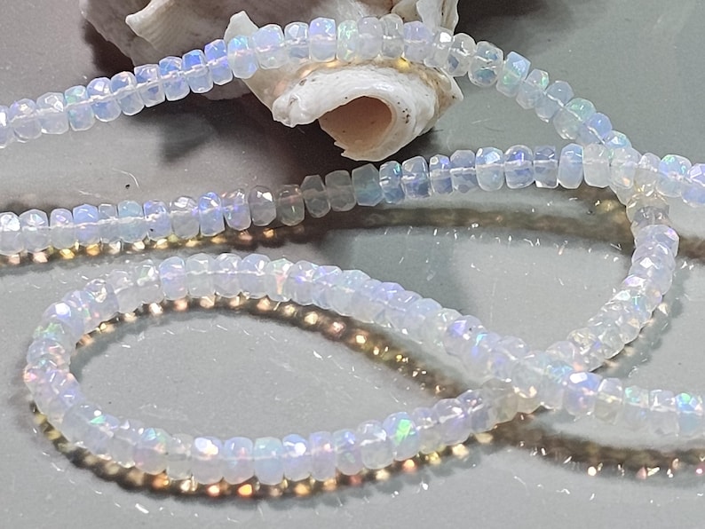May include: A strand of small, faceted, translucent white opal beads with iridescent flashes of blue, green, and yellow light. The beads are circular and strung together in a continuous line, displaying a reflective, polished surface texture.