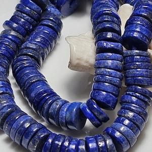 Puede incluir: Primer plano de un collar hecho de cuentas en forma de disco de color azul oscuro. Las cuentas tienen una superficie pulida y están ensartadas. El collar se muestra sobre una superficie blanca, con una concha marina visible en el fondo. Las cuentas son de un azul intenso.