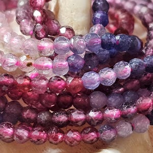 Op de afbeelding: Een verzameling gefacetteerde, ronde edelsteenkralen in verschillende tinten dieppaars, magenta en doorschijnend roze. De kleine, gepolijste kralen zijn gerangschikt in meerdere strengen en tonen een reflecterend kleurverloop.