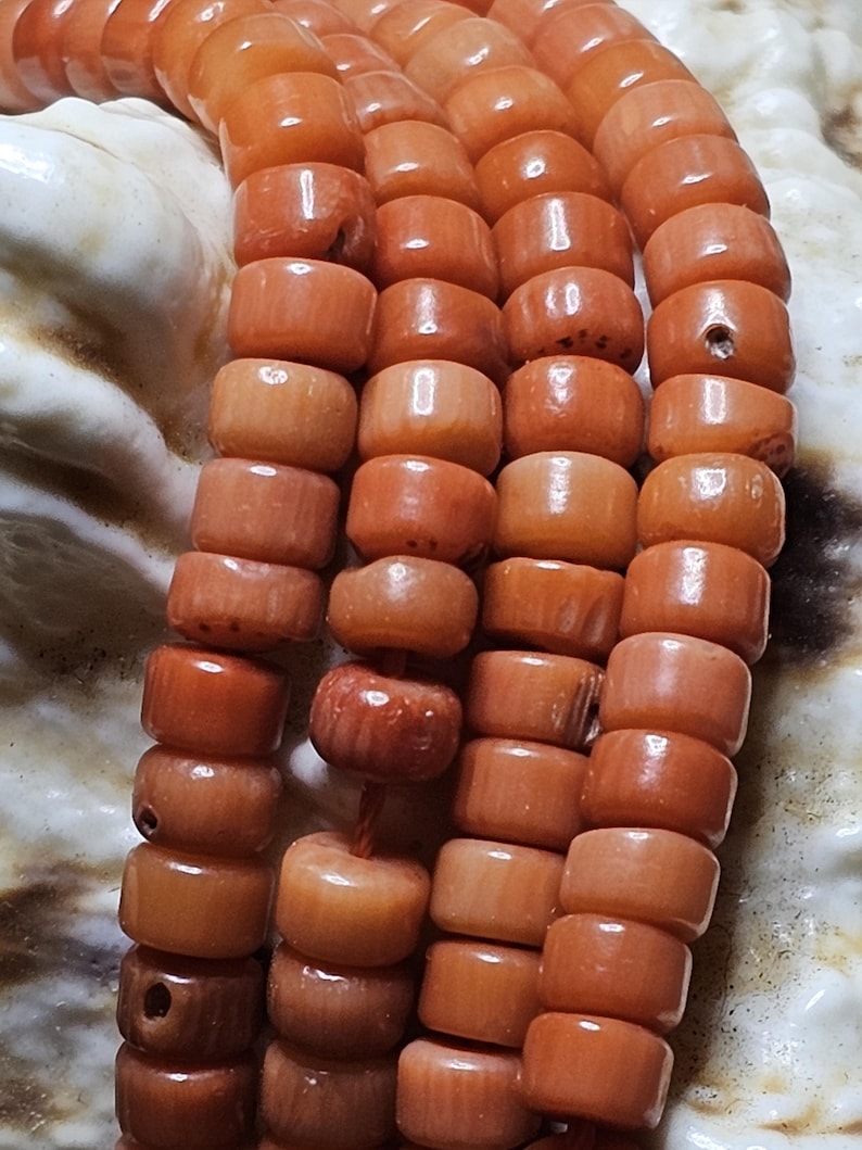 Op de afbeelding: Close-up van een koraal ketting. De ketting heeft meerdere strengen cilindrische, rood-oranje koraal kralen. De kralen zijn aan elkaar geregen, waarbij hun natuurlijke kleur en textuur worden getoond. De achtergrond is een vage, gebroken witte ondergrond.