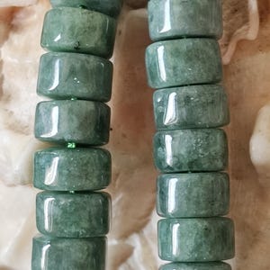 May include: A close-up of a necklace featuring cylindrical, jade-green beads. The beads are strung together with a green cord. The beads have a polished, glossy finish, reflecting light. The necklace is likely a piece of jewelry.