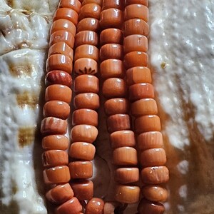 Op de afbeelding: Close-up van een ketting gemaakt van oranje, cilindrische kralen. De kralen zijn aan elkaar geregen, waardoor een gelaagd effect ontstaat. De ketting is geplaatst tegen een getextureerde, gebroken witte achtergrond, mogelijk een zeeschelp.
