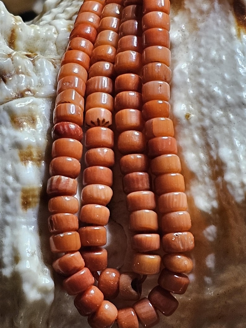 Op de afbeelding: Close-up van een ketting gemaakt van oranje, cilindrische kralen. De kralen zijn aan elkaar geregen, waardoor een gelaagd effect ontstaat. De ketting is geplaatst tegen een getextureerde, gebroken witte achtergrond, mogelijk een zeeschelp.