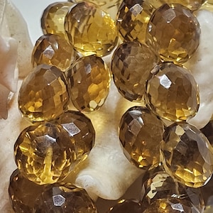 May include: Close-up of faceted, oval-shaped citrine gemstones. The gems are a warm, amber color with multiple facets that catch the light. The citrines are resting on a white, textured surface, creating a contrast in color and texture.