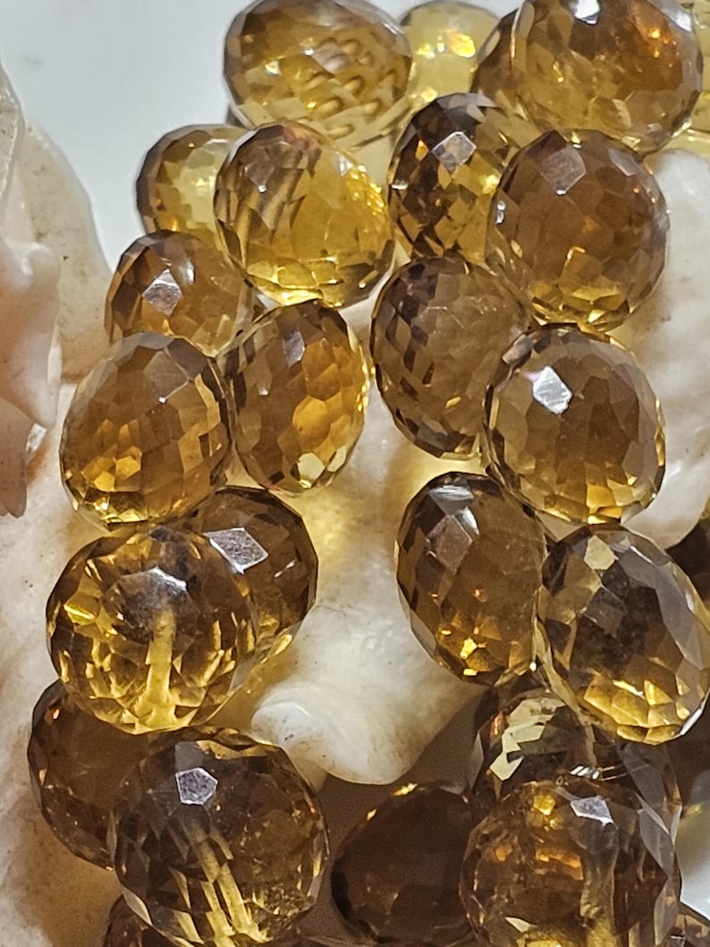 May include: Close-up of faceted, oval-shaped citrine gemstones. The gems are a warm, amber color with multiple facets that catch the light. The citrines are resting on a white, textured surface, creating a contrast in color and texture.