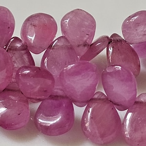 May include: A close-up of a strand of pink gemstone beads. The beads are smooth and polished, with a teardrop shape. They are arranged in a random pattern.