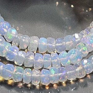 May include: A strand of faceted, translucent white beads with an iridescent, opalescent sheen. Each bead features a circular, flattened disc shape with multiple cut surfaces that reflect light, creating flashes of blue, green, and pink colour.
