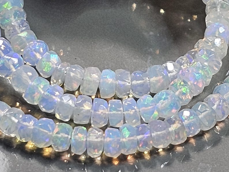 May include: A strand of faceted, translucent white beads with an iridescent, opalescent sheen. Each bead features a circular, flattened disc shape with multiple cut surfaces that reflect light, creating flashes of blue, green, and pink colour.