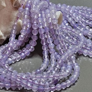Puede incluir: Primer plano de varios hilos de cuentas facetadas de color púrpura claro. Las cuentas son pequeñas y redondas, con una calidad brillante y translúcida. Los hilos están drapeados sobre una superficie gris, con una concha en el fondo.