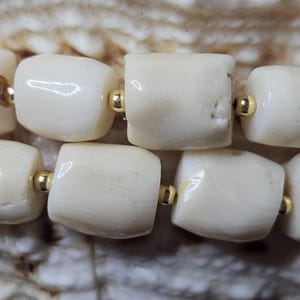 May include: Close-up of a strand of off-white, barrel-shaped beads with gold-colored spacers. The beads have a natural, organic texture and are strung together, creating a simple yet elegant design. The background is a textured, light brown surface.