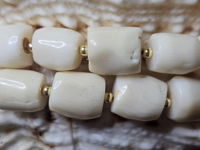 May include: Close-up of a strand of off-white, barrel-shaped beads with gold-colored spacers. The beads have a natural, organic texture and are strung together, creating a simple yet elegant design. The background is a textured, light brown surface.