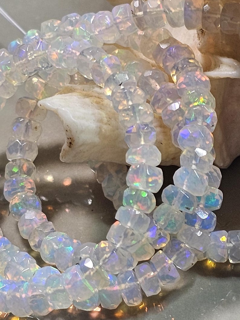 May include: A strand of faceted, translucent white beads with an iridescent finish that displays flashes of green, blue, and orange light. The beads are round and disc-shaped, strung together in a continuous line, reflecting light across their surfaces.