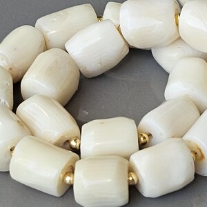 May include: Close-up of a necklace featuring large, off-white, barrel-shaped beads. The beads are strung together with gold-colored spacers. The necklace is displayed on a gray surface.