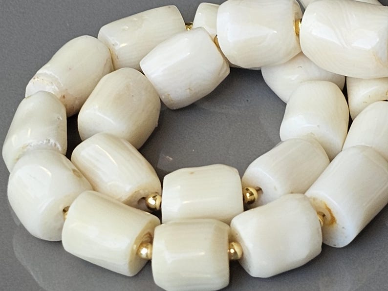May include: Close-up of a necklace featuring large, off-white, barrel-shaped beads. The beads are strung together with gold-colored spacers. The necklace is displayed on a gray surface.