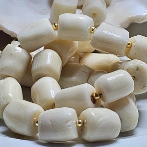May include: Close-up of a necklace featuring large, off-white, barrel-shaped beads. The beads are strung together with gold-colored accents. The necklace is displayed on a white surface, showcasing the natural variations in the beads.
