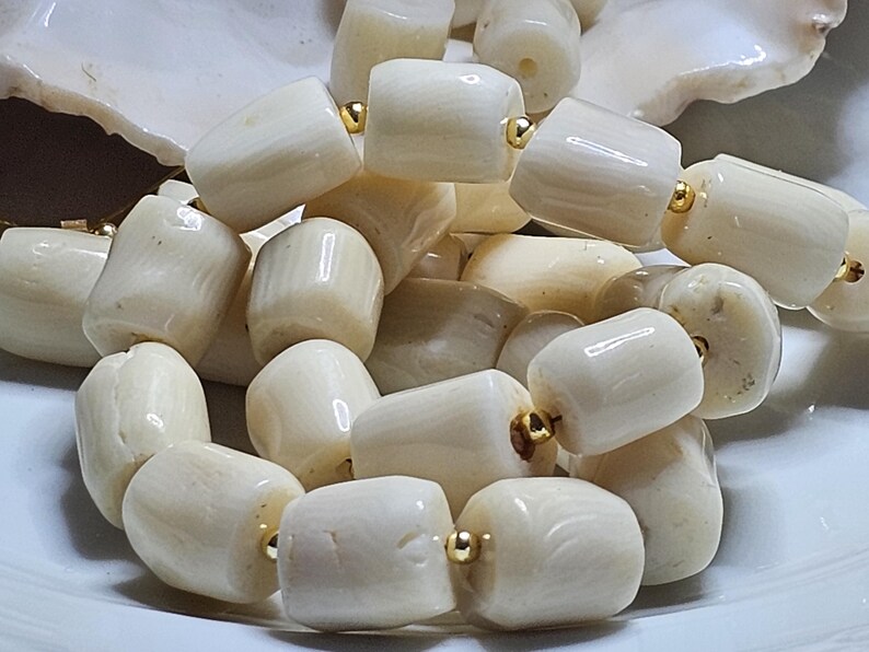 May include: Close-up of a necklace featuring large, off-white, barrel-shaped beads. The beads are strung together with gold-colored accents. The necklace is displayed on a white surface, showcasing the natural variations in the beads.