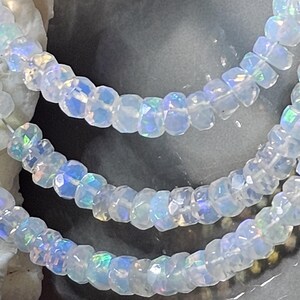 May include: A strand of faceted, translucent white beads with an iridescent, opalescent sheen. The small, circular beads are strung together, displaying subtle flashes of blue, green, and pink light across their polished, multi-faceted surfaces.