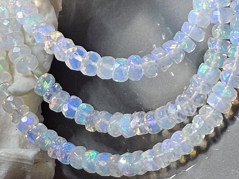 May include: A strand of faceted, translucent white beads with an iridescent, opalescent sheen. The small, circular beads are strung together, displaying subtle flashes of blue, green, and pink light across their polished, multi-faceted surfaces.