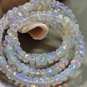 May include: A strand of faceted, translucent white opal beads arranged in a circular coil. Each small, round bead features a polished surface that reflects iridescent flashes of blue, green, and pink light throughout the gemstone strand.