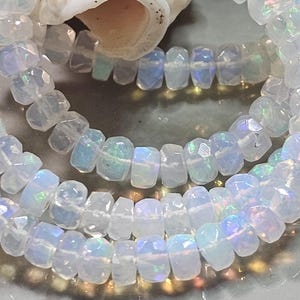 May include: A strand of faceted, translucent white opal beads with iridescent flashes of blue, green, and pink. The circular, disc-shaped beads are strung together on a thin, clear filament, creating a reflective, multi-layered appearance.