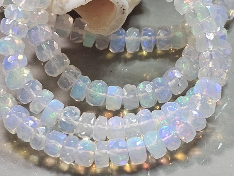 May include: A strand of faceted, translucent white opal beads with iridescent flashes of blue, green, and pink. The circular, disc-shaped beads are strung together on a thin, clear filament, creating a reflective, multi-layered appearance.