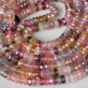 Könnte beinhalten: Ein Strang aus facettierten Rondellperlen in verschiedenen durchscheinenden Farben wie Rosa, Gelb, Grün und Blau. Die kleinen, kreisförmigen Perlen sind in einem gewundenen Muster angeordnet und zeigen eine reflektierende, polierte Oberfläche.