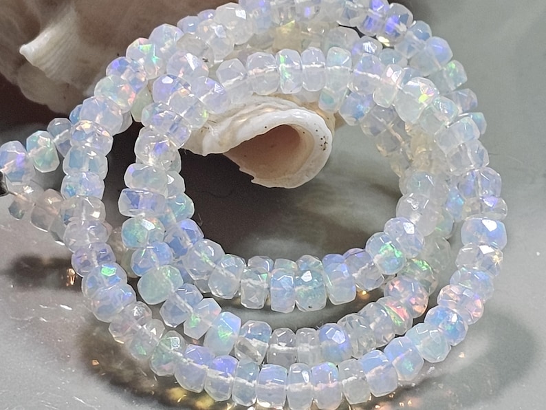 May include: A strand of faceted, translucent white beads with an iridescent finish that reflects flashes of blue, green, and pink light. The small, circular beads are strung together in a loose coil, showcasing their reflective, multi-faceted surfaces.